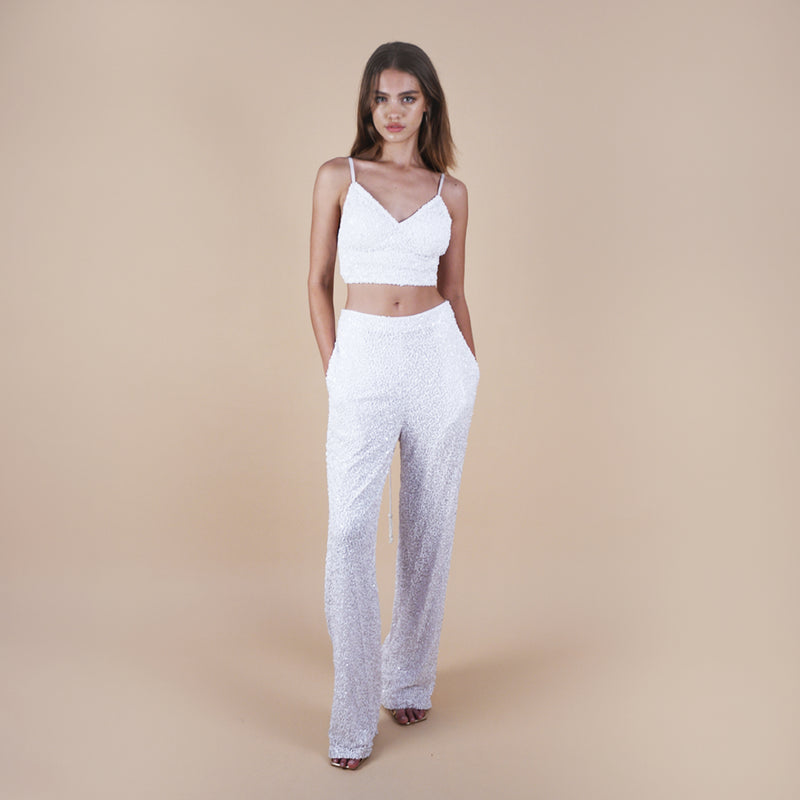off-white Anastasia Beaded Bralette Crop Top. Handcrafted with intricate beadwork, this versatile party outfit features a lace-up tie closure at the back, ensuring a perfect fit. Stand out at events with this chic and stylish crop top. Front view on the model with sequin pants showing pockets